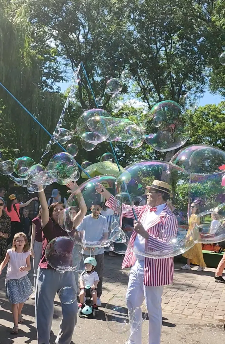 Dans un parc, Rigadin en veste rayée et canotier lance des bulles géantes au-dessus des enfants