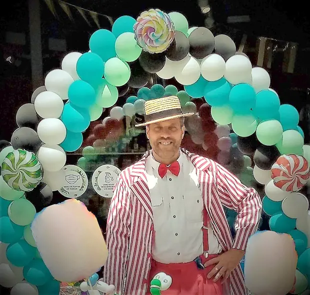 Rigadin en canotier et veste rayée, une barbe à papa dans chaque main, devant une arche de ballons