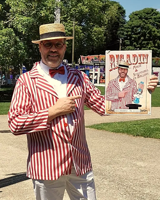 Rigadin en canotier et veste rayée rouge et blanc, dans un parc, montre son affiche « Rigadin, magie et sculptures en ballons »