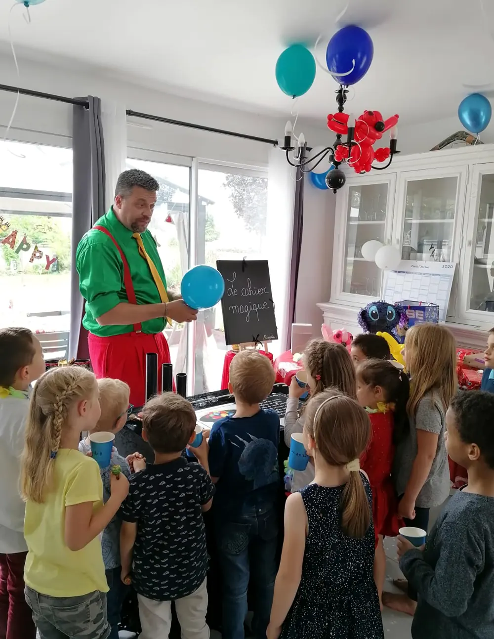 Dans un salon décoré de ballons, Rigadin sculpte un ballon devant un groupe d’enfants
